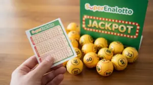 Biglietto del SuperEnalotto tenuto in mano con sfondo di palline numerate e cartello con scritta Jackpot