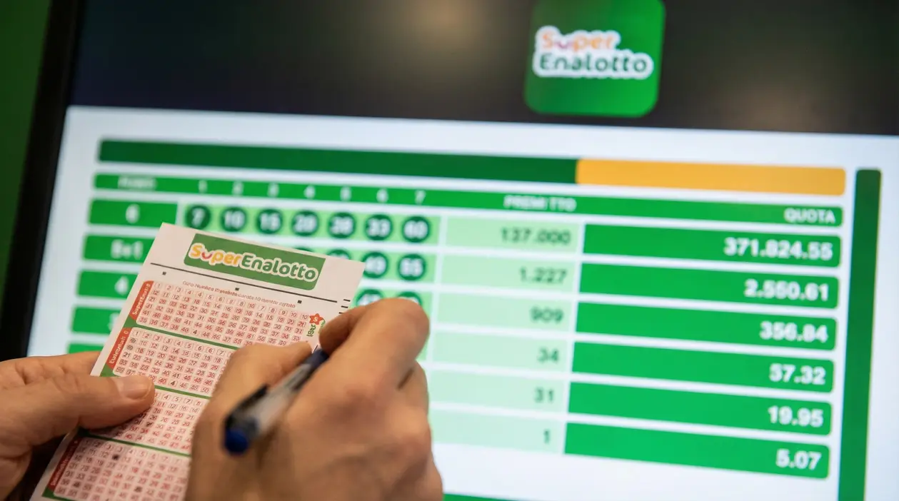 Una persona compila una schedina del Superenalotto davanti a uno schermo con i risultati